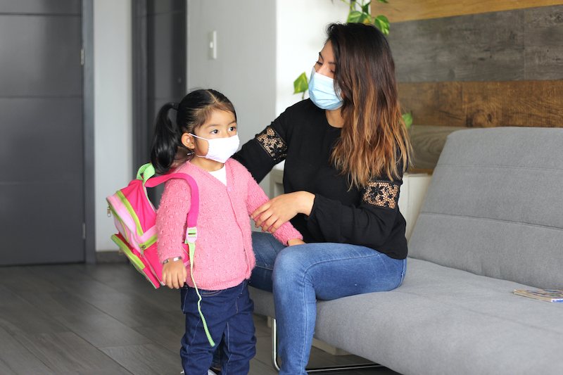 Mother wearing medical mask helping daughter wearing medical mask with her backpack